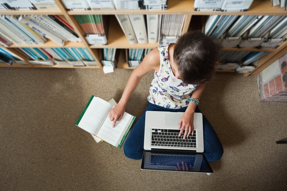 High angle view of a female student sitting against bookshelf with laptop on the library floor-1-1